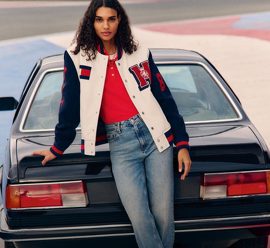 imagen de una mujer con chamarra blanca con azul y blusa roja recargada en un auto mobil negro. TOMMY HILFIGER