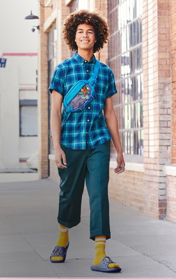 Hombre camiando en la calle, viste camusa a cuadros de color azul, pantalon tipo pescador color verde, calcetas amarillas y unmos CROCS de color morado. CROCS