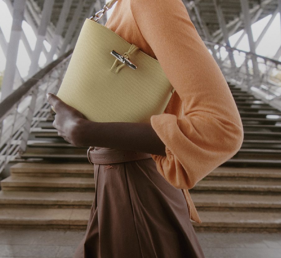 Imagen de mujer con bolso verde, blusa naranja y falda cafe. LONGCHAMP.