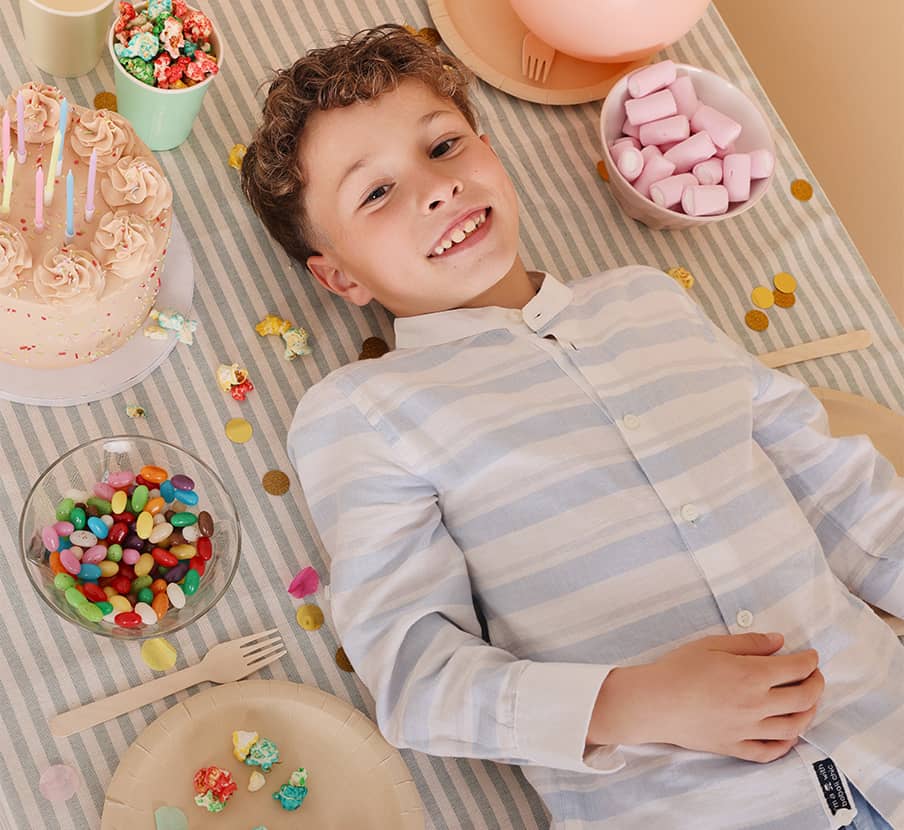 Imagen de un niño acostado en una manta, al rededor de el muchos dulces y un pastel, el niño viste una camisa de color blanca con rayas azul claro de la marca BOBOLI