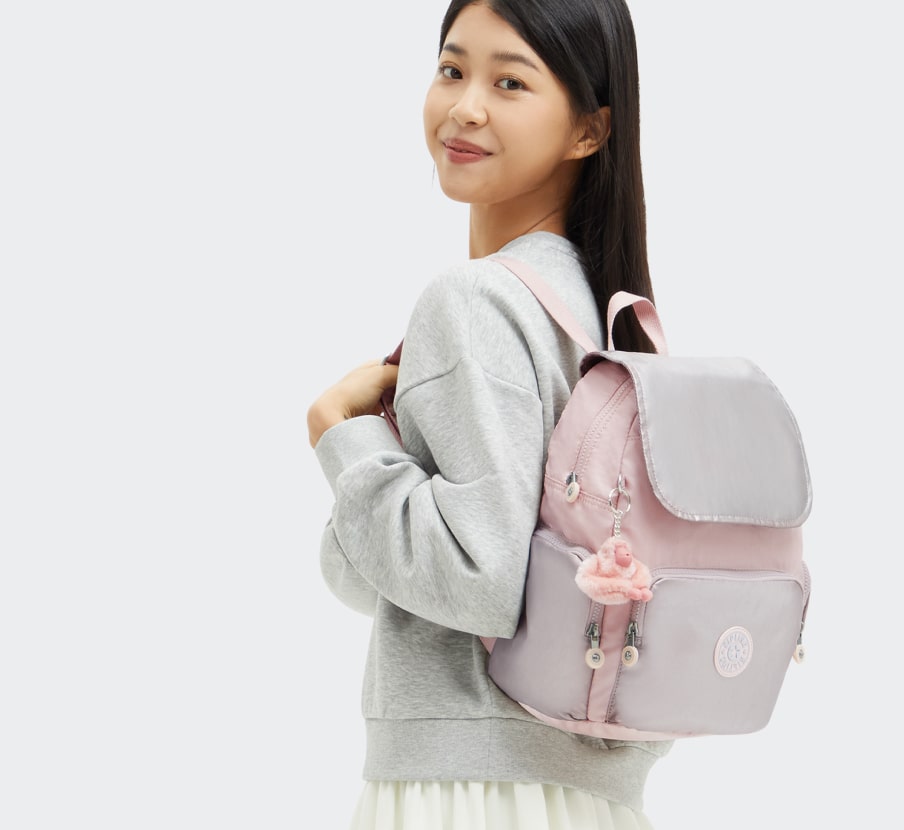 imagen de niña con sueter gris y falda blanca llevando mochila rosa con gris, KIPLING