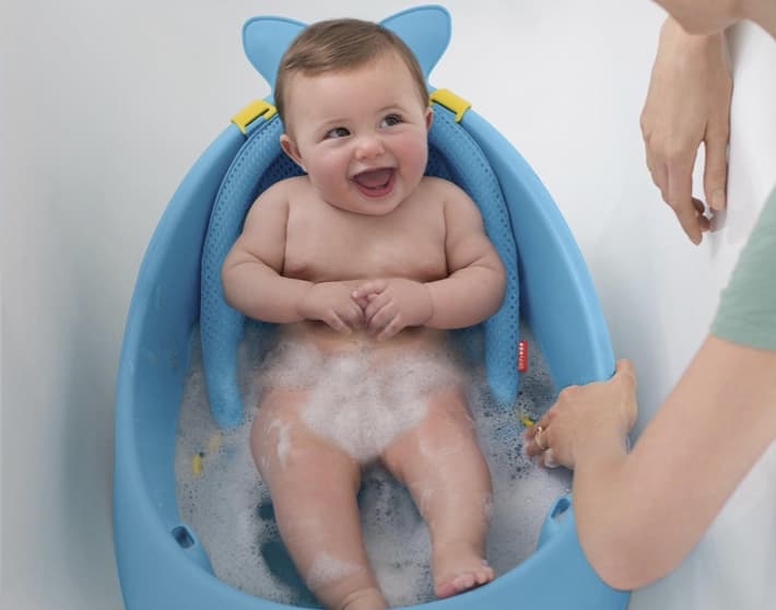 Imagen de un bebe al cual lo estan bañando en una bañera color azul de la marca, SKIP HOP