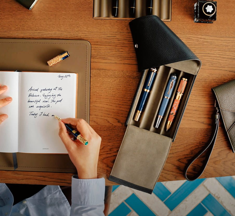 Imagen de un un juego de plumas de la marca MONTBLANC, junto a ellas una mano escribiendo sobre un cuaderno, MONTBLANC