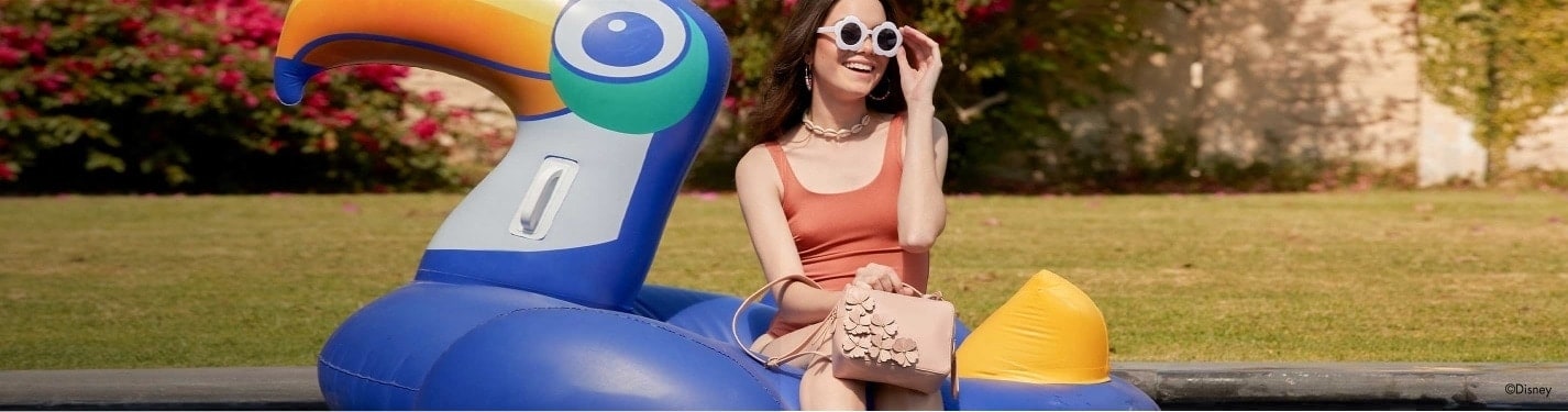 Mujer sentada en un inflable para alberca con forma de pelicano color azul, viste un traje de baño de una pieza color marron, y lleva un bolso color beige, W CAPSULE
